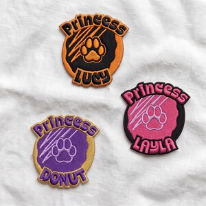 May include: Three circular embroidered patches featuring a central paw print with claw marks. One patch is orange and black with the text Princess Lucy, one is pink and black with Princess Layla, and one is purple and gold with Princess Donut.