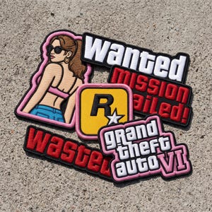 May include: A collection of embroidered patches featuring a woman in sunglasses, a yellow square with a black R and white star, and text patches reading Wanted, Mission Failed, Wasted, and Grand Theft Auto VI in various colors including pink, red, and white.