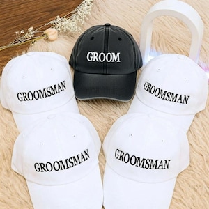 May include: A collection of baseball caps, including one black cap with the word "Groom" in white, surrounded by white caps with "Groomsman" in black. The caps are arranged on a textured surface, suggesting a wedding theme.