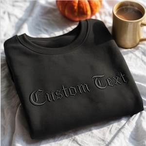 May include: Black crewneck sweatshirt with the embroidered text "Custom Text" in a gothic font. The sweatshirt is displayed on a white surface with a small pumpkin and a mug of coffee in the background.
