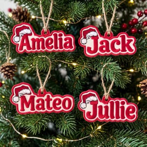 May include: Four custom Christmas ornaments featuring the names Amelia, Jack, Mateo, and Jullie in red lettering with white outlines. Each ornament includes a small red and white Santa hat graphic and hangs from a natural jute twine loop.