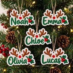 May include: Five white Christmas ornaments featuring brown reindeer antlers, green holly leaves, red berries, and the names Noah, Emma, Chloe, Olivia, and Lucas embroidered in green. Each ornament hangs from a thin jute twine string.