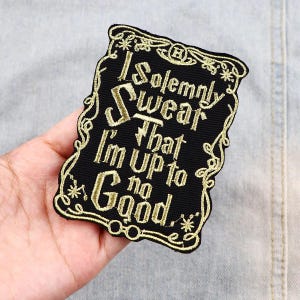 May include: Black embroidered patch with gold text that reads "I Solemnly Swear That I'm Up to No Good." The patch has a decorative border and is held in a hand. The patch is likely for clothing or accessories.