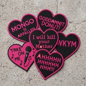 May include: A collection of six heart-shaped embroidered patches with black fabric and bright pink borders. The patches feature text including "MONGO is APPALLED," "GODDAMNIT DONUT!," "I will kill your Mother," "WKYM," "TAKE ON ME" with a microphone, and "AHHHHHHHHHHH!!!."