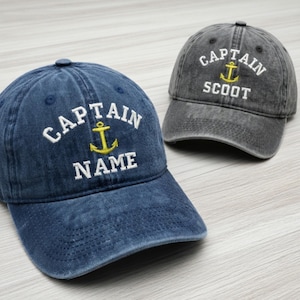 May include: Two baseball caps, one denim blue and one gray, with the word "CAPTAIN" arched above an anchor graphic. The blue cap has the word "NAME" below the anchor, while the gray cap says "SCOOT". Both caps have a curved brim.