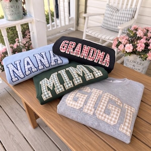 May include: Four crewneck sweatshirts featuring collegiate-style block lettering with gingham patterns. The light blue sweatshirt says NANA, the black one says GRANDMA, the dark green one says MIMI, and the heather gray one says GIGI.