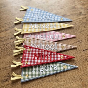 May include: A set of six triangular pennants featuring a gingham check pattern in blue, yellow, pink, tan, red, and green. Each pennant has a white embroidered word including Tigers, Mascot, Custom, School, Nini, and Tigers, with two gold stars.