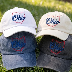 May include: Four baseball caps in white, cream, blue denim, and black denim. Each cap features an embroidered red outline of the state of Ohio with the word Ohio stitched in blue script lettering across the center of the front panel.