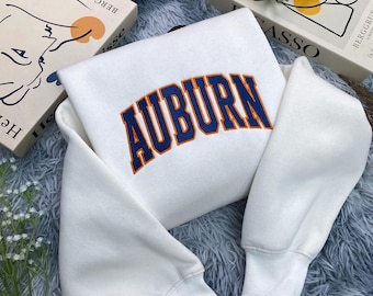 May include: A white sweatshirt with the word "AUBURN" in large, arched, navy blue letters with orange trim. The sweatshirt has long sleeves with ribbed cuffs. The background includes books and decorative elements.