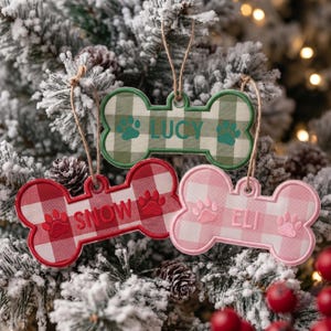 May include: Three bone-shaped ornaments featuring a buffalo plaid pattern. The top ornament is green with the name LUCY and paw prints. The bottom left is red with the name SNOW and paw prints. The bottom right is pink with the name ELI and paw prints.
