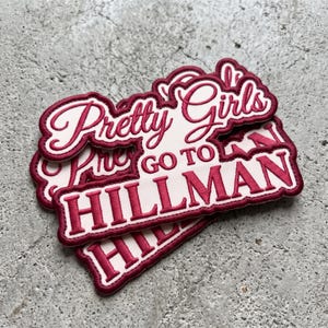 May include: A stack of embroidered patches featuring a white background with dark red borders and lettering. The text reads Pretty Girls GO TO HILLMAN in a combination of cursive and block fonts. The patches have a stitched edge finish.