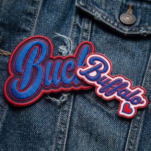 May include: Two embroidered patches attached to a blue denim jacket. The larger patch features the word Buck in blue script with a red border. The smaller patch displays the word Buffalo in blue script with a white background and red border, ending with a red heart.