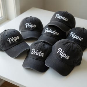 May include: A collection of dark gray baseball caps with white embroidered text. The caps feature various names like "Pôpo", "Pāpaw", "Dāda", "Pāpa", and "Grandpa". The caps are arranged on a white surface.
