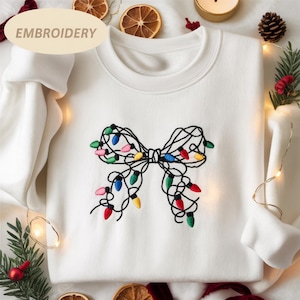 May include: White sweatshirt with an embroidered bow design. The bow is made of black thread and features colorful Christmas lights in red, green, blue, and yellow. The word "EMBROIDERY" is printed on the top left.