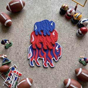 May include: A stack of three buffalo-shaped patches in blue and red, surrounded by miniature American footballs, small football player figurines, a yellow goal post, and several small flag markers on a textured gray surface.
