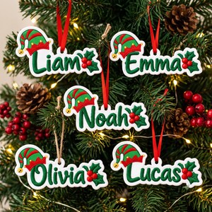 May include: Five white Christmas ornaments featuring the names Liam, Emma, Noah, Olivia, and Lucas in green script. Each ornament is decorated with a red and green elf hat and a cluster of red holly berries, suspended by red ribbons.