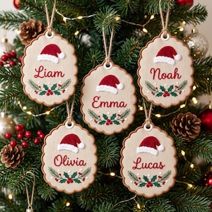 May include: Five scalloped wooden ornaments hanging by twine. Each features a red and white Santa hat, a green holly sprig with red berries, and a name embroidered in red script: Liam, Emma, Noah, Olivia, and Lucas.
