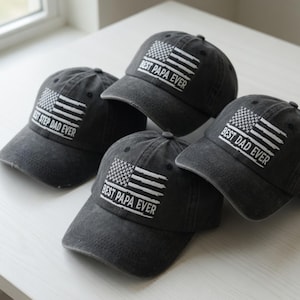 May include: Four charcoal gray baseball caps with a distressed American flag design. Each cap features the text "Best Papa Ever" or "Best Dad Ever" or "Best Step Dad Ever" in white block letters. The caps are arranged on a white surface.