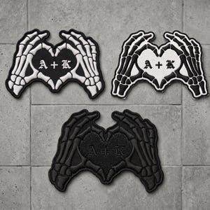 May include: Three embroidered patches featuring skeleton hands forming a heart shape. One patch has white bones on a black background, one has black bones on a white background, and one is entirely black. Each heart contains the text A + K.