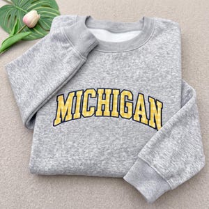 May include: A heather gray crewneck sweatshirt featuring the word MICHIGAN in a yellow and white gingham pattern with a dark blue outline. The long-sleeved garment has ribbed cuffs and a ribbed neckline, showcasing a classic athletic design.