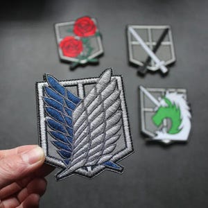 May include: Embroidered patches featuring various designs. One patch displays blue and white wings, while others showcase a rose, a sword, and a green unicorn. The patches are gray, white, blue, and green.