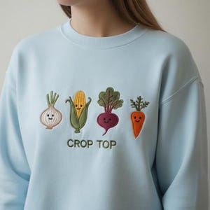 May include: Light blue sweatshirt with embroidered vegetables: an onion, corn, beet, and carrot. The text "CROP TOP" is embroidered below the vegetables. The sweatshirt is a solid light blue color.