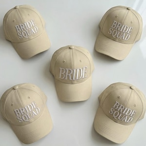 May include: Five beige baseball caps. Four caps are embroidered with "BRIDE SQUAD" in white, and one cap is embroidered with "BRIDE" in white. The caps are arranged on a light-colored surface.