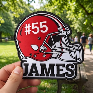 May include: A red and white embroidered patch featuring a football helmet with the number 55 printed in white on the side. Below the helmet, the name JAMES is embroidered in bold black capital letters with a white border on a black background.