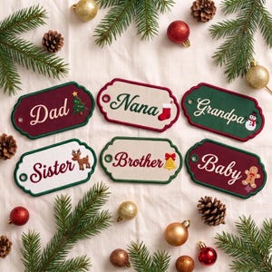 May include: Six rectangular holiday gift tags with scalloped edges, featuring embroidered names and festive icons. The tags are labeled Dad, Nana, Grandpa, Sister, Brother, and Baby, displayed against a background of pine branches and ornaments.