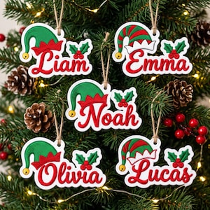 May include: Five white Christmas ornaments featuring red cursive names Liam, Emma, Noah, Olivia, and Lucas. Each ornament displays a green and red elf hat with a yellow bell and a sprig of green holly leaves with red berries, hanging by twine.