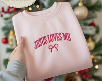 May include: A light pink sweatshirt with the embroidered text "JESUS LOVES ME" in a cursive font, with a small bow beneath. The word "EMBROIDERY" is in a white oval in the upper left corner. The sweatshirt is folded.