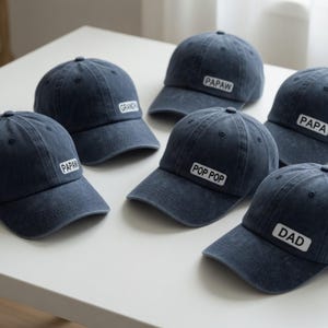 May include: A collection of navy blue baseball caps with white rectangular patches. Each patch features a different word: PAPAW, GRANDPA, PAPA, POP POP, and DAD. The caps are arranged on a white surface.