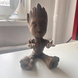 May include: A brown figurine of Groot from Guardians of the Galaxy. The figure is in a seated position with arms outstretched. The figurine has a detailed, textured appearance, with a small face and large feet.