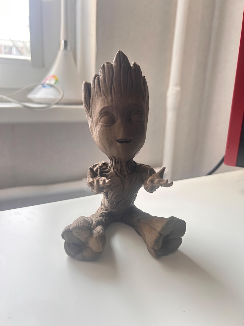 以下が含まれることがあります： ガーディアンズ・オブ・ギャラクシーのグルートの茶色のフィギュア。腕を広げた座った姿勢です。小さな顔と大きな足があり、詳細で質感のある外観をしています。