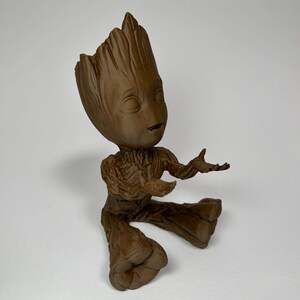 以下が含まれることがあります： ガーディアンズ・オブ・ギャラクシーのキャラクター、グルートの茶色のフィギュア。腕を広げた座った姿勢です。フィギュアは詳細で質感のある外観で、笑顔の表情をしています。