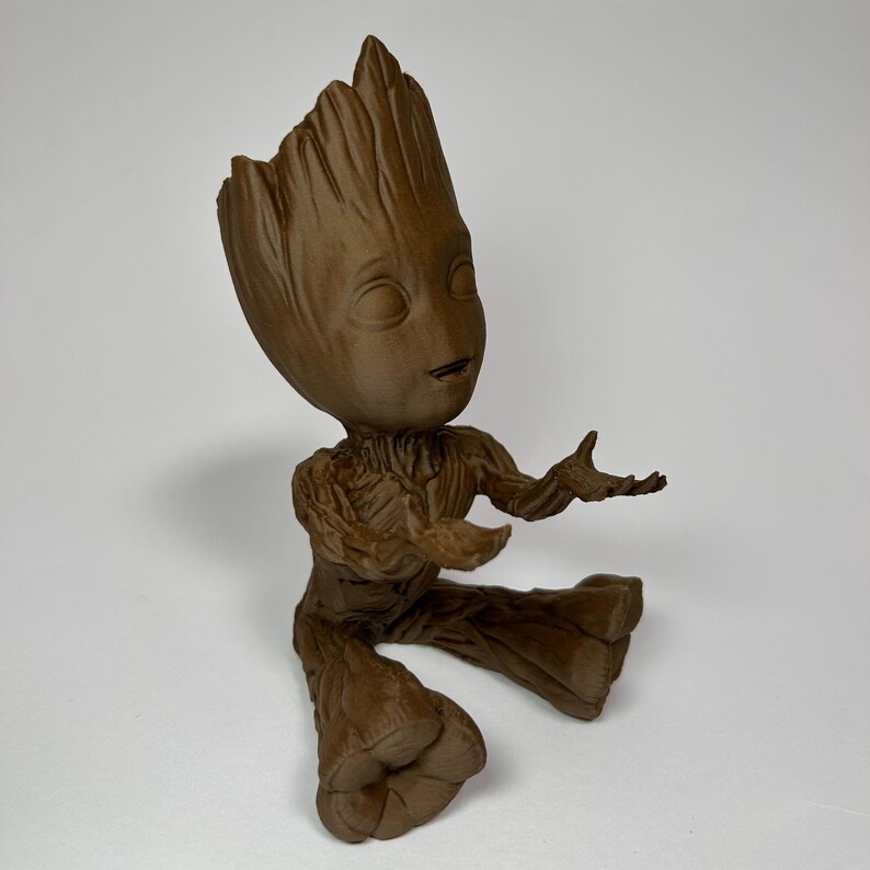 以下が含まれることがあります： ガーディアンズ・オブ・ギャラクシーのキャラクター、グルートの茶色のフィギュア。腕を広げた座った姿勢です。フィギュアは詳細で質感のある外観で、笑顔の表情をしています。