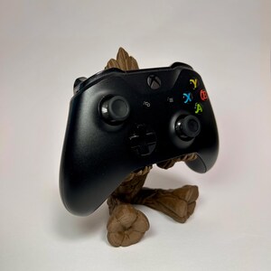 以下が含まれることがあります： 黒いXboxコントローラーが、木目調の茶色のスタンドに置かれています。コントローラーにはカラーボタンと2つのジョイスティックがあります。スタンドは人気映画のキャラクターの形をしています。