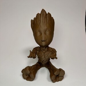 以下が含まれることがあります： ガーディアンズ・オブ・ギャラクシーシリーズのキャラクター、グルートの茶色のフィギュア。腕を広げ、笑顔で座っている姿です。フィギュアの高さは約15cmです。