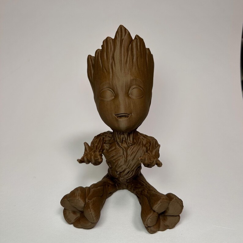 以下が含まれることがあります： ガーディアンズ・オブ・ギャラクシーシリーズのキャラクター、グルートの茶色のフィギュア。腕を広げ、笑顔で座っている姿です。フィギュアの高さは約15cmです。