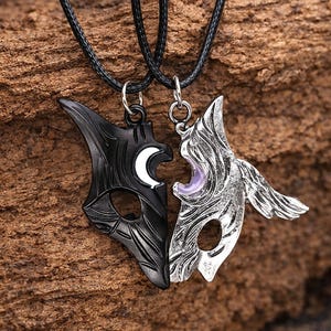 May include: Two matching pendant necklaces. One pendant is black with a white crescent moon, the other is silver with a purple gem. Both pendants are shaped like stylized masks and are connected to black cords.