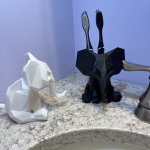 May include: A white geometric elephant figurine and a black elephant-shaped toothbrush holder with two toothbrushes. The items are on a speckled countertop near a bathroom sink.