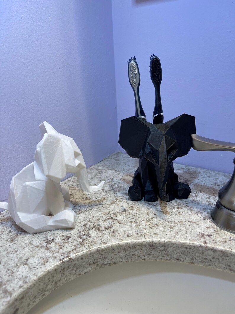 May include: A white geometric elephant figurine and a black elephant-shaped toothbrush holder with two toothbrushes. The items are on a speckled countertop near a bathroom sink.