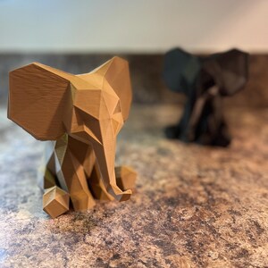 May include: Two geometric elephant figurines. The foreground elephant is light brown, while the background elephant is black. Both are sitting with their trunks down. The figurines have a modern, angular design.
