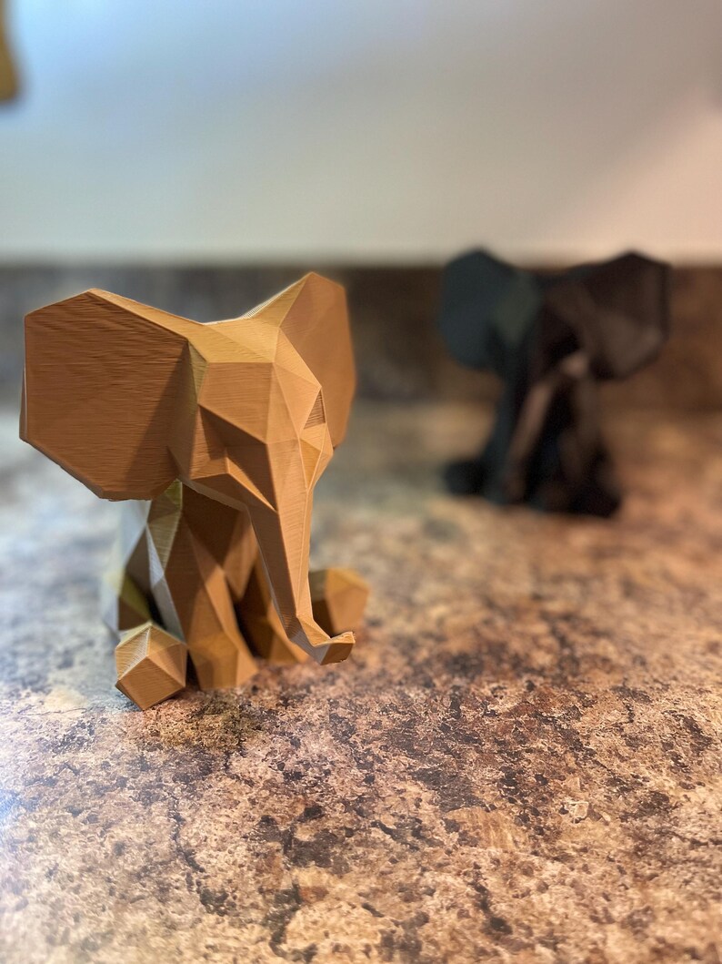 May include: Two geometric elephant figurines. The foreground elephant is light brown, while the background elephant is black. Both are sitting with their trunks down. The figurines have a modern, angular design.