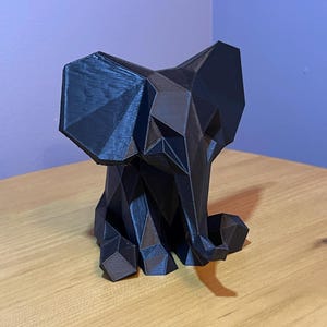 May include: A black, geometric elephant sculpture. The elephant is made of many flat, triangular surfaces, giving it a faceted appearance. The sculpture is sitting on a wooden surface.