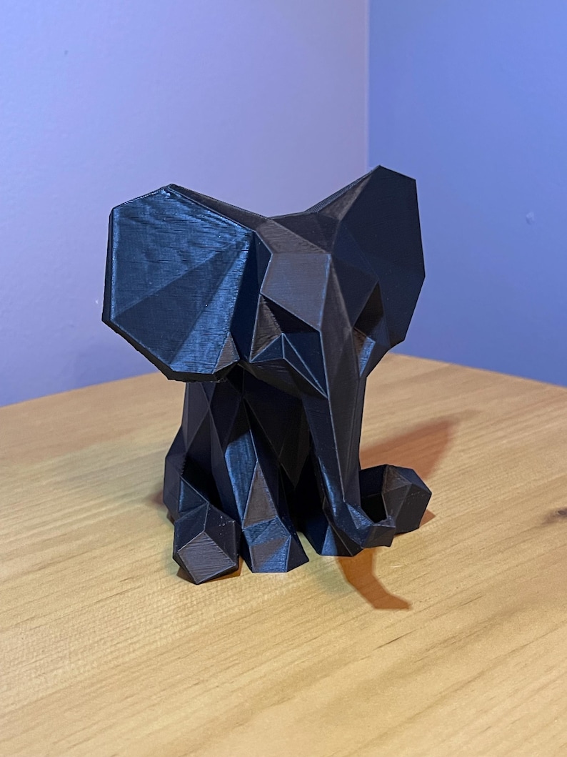 May include: A black, geometric elephant sculpture. The elephant is made of many flat, triangular surfaces, giving it a faceted appearance. The sculpture is sitting on a wooden surface.