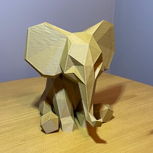 May include: A geometric, light brown elephant sculpture. The elephant is sitting with its ears spread and trunk down. The sculpture is made of many flat, angular planes, giving it a modern, polygonal look. The elephant is on a wooden surface.