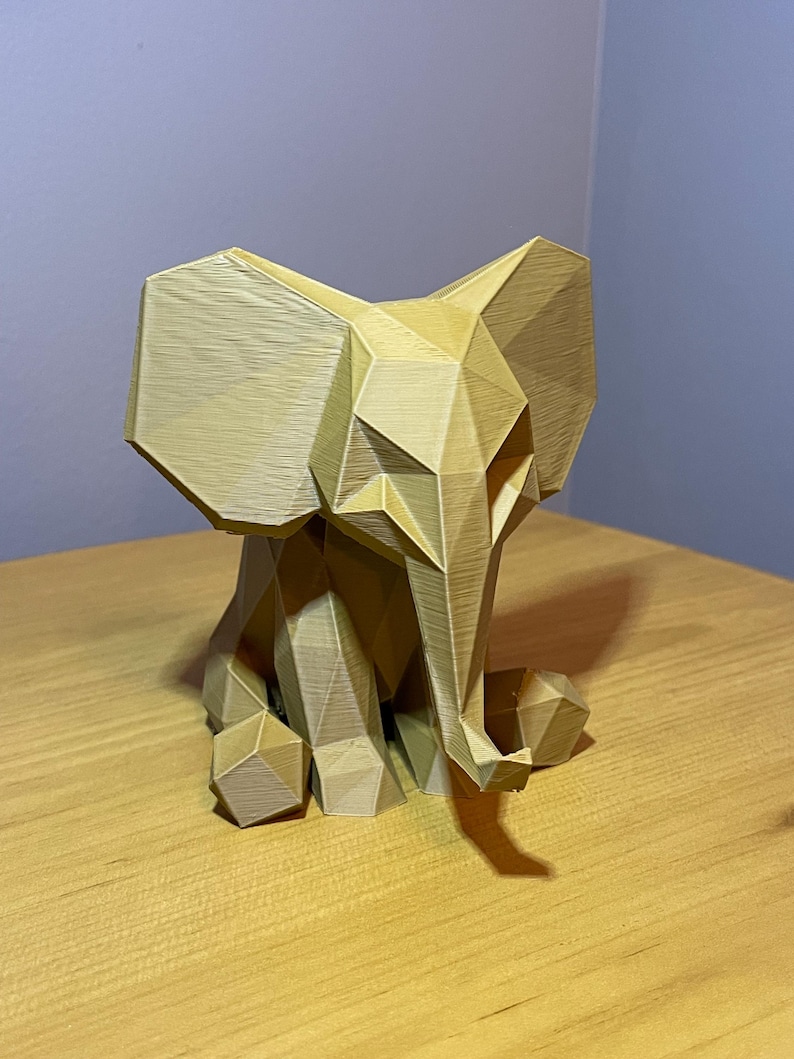 May include: A geometric, light brown elephant sculpture. The elephant is sitting with its ears spread and trunk down. The sculpture is made of many flat, angular planes, giving it a modern, polygonal look. The elephant is on a wooden surface.
