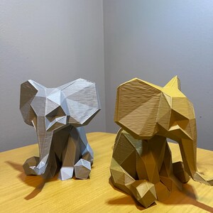 May include: Two geometric elephant figurines. One is silver, the other is gold. Both are sitting with their trunks down and ears out. The figurines are on a light brown wooden surface.
