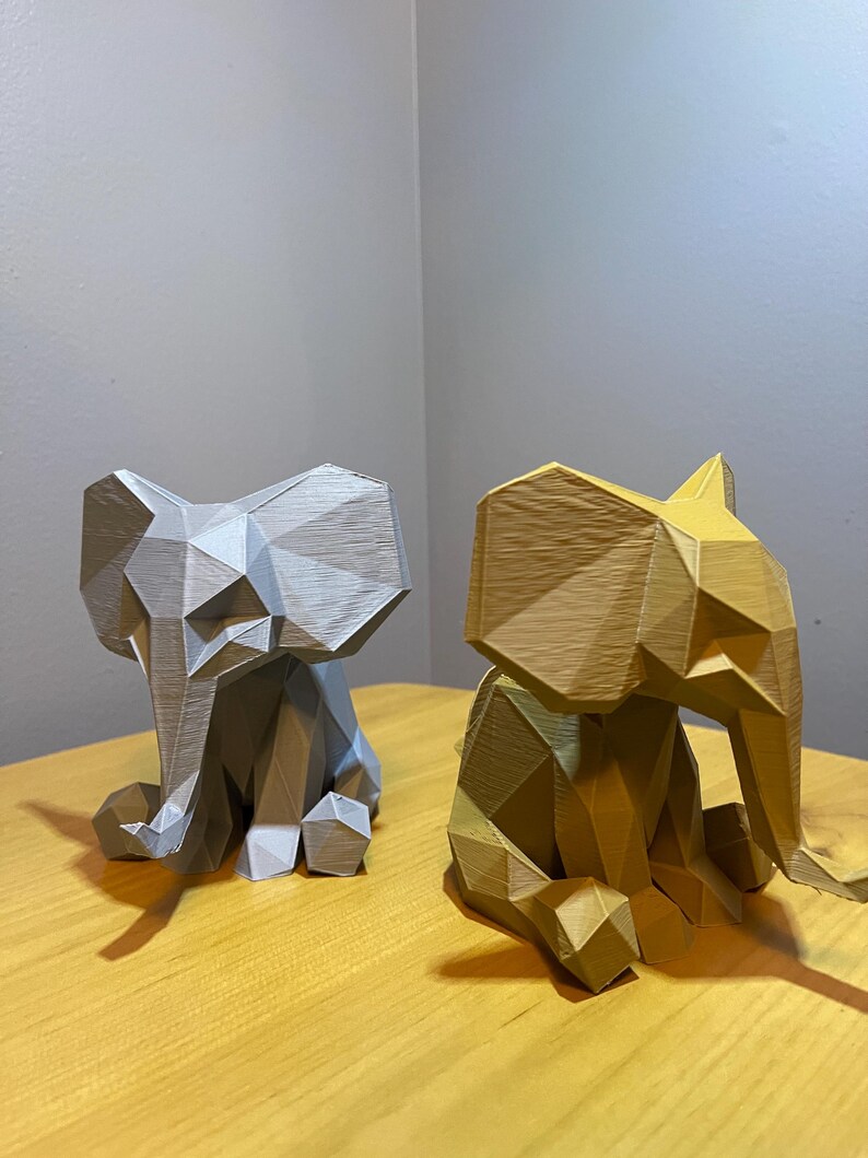 May include: Two geometric elephant figurines. One is silver, the other is gold. Both are sitting with their trunks down and ears out. The figurines are on a light brown wooden surface.